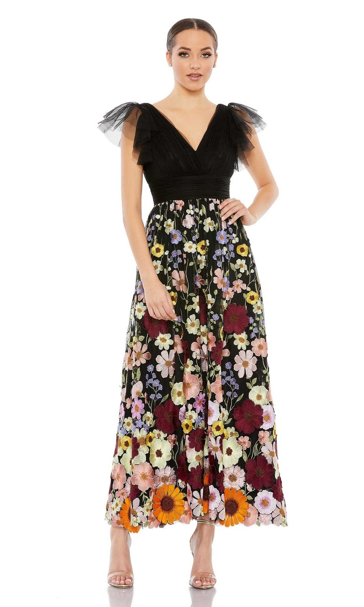 A woman wears the Mac Duggal 68204 Tea-Length Floral Formal Dress, featuring tulle shoulders and a sleeveless black top with a floral-patterned skirt, standing against a white background.